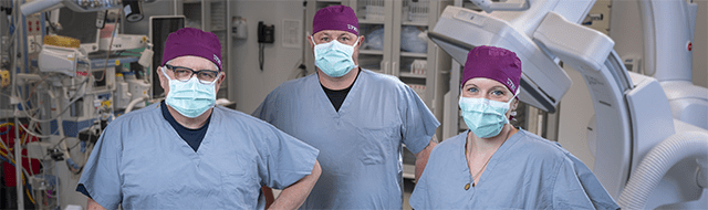Three Minimally Invasive Gynecologic Surgeons standing in a operating room wearing blue  scrubs and purple surgical caps. 