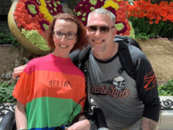 Jessica sits in her wheelchair in front of a garden with a floral sculpture in the shape of a butterfly. Her husband James hugs her. She wears a bright t-shirt with red, orange, green and blue stripes.