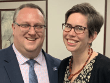 Merissa wears a black and white floral print dress and a black cardigan. She has red earrings and a red necklace. She has short brown hair and wears glasses. She stands with her husband who wears a blue suit and blue tie.