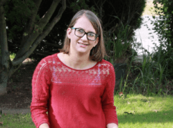 Sarah Meyers sits outside on a hay bale next to her small black and white dog. She wears a red sweater, jeans, and brown ugg boots. She has brown hair and glasses. There are two pumpkins resting by her feet.