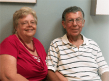 Earl and his wife sit in a doctor's office. He wears a black and white striped polo shirt. She wears a coral blouse and beaded necklace. He has gray hair. They both wear glasses.