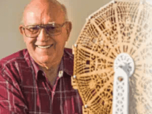 John smiles. He wears tinted yellow glasses and a red and white plaid t-shirt. He is bald. He shows a 3-d model of a ferris wheel.