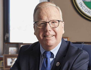 Mark sits in his office. He has rounded glasses, and wears a black suit with a blue tie. He has white hair cropped short.