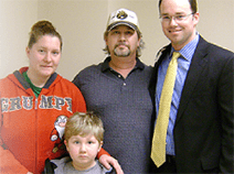 William stands with his family and his doctor. He wears a gray shirt and a baseball cap. His wife wears an orange sweatshirt, and their son wears a gray hoodie. The doctor is wearing a suit. 