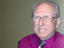 Gene has gray hair and wears glasses. He is wearing a purple collared button down shirt and a shiny, silver tie.