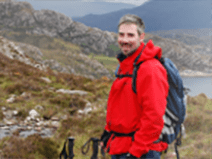 Richard is in a valley surrounded by mountains. He wears a weatherproof jacket in red, and jeans. He has gloves and a hiking backpack, as well as trekking poles.