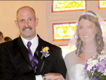 David Smith walking the bride down the aisle. He wears a suit with a yellow flower and a purple tie. He has a moustache and is bald. He looks very happy.