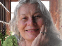 A woman is resting her chin on her hand. She wears two rings on her ring and pinky finger. She has long silver hair. She wears a green blouse that has a flower trim.