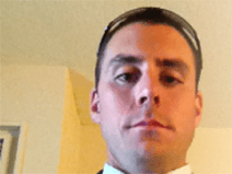 He has short brown hair. He wears a black suit with a white tuxedo shirt and a white tie.