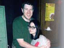 A man in a green shirt poses for a picture hugging a woman. He wears a green t-shirt and jeans. He has brown hair. She has black hair, glasses, and wears a white, red, and yellow hoodie.