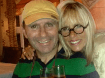 He wears a polo shirt that has green and black stripes. He has on a yellow baseball cap. He is smiling. A woman with round glasses and blonde hair poses with him for the picture.
