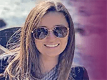 A young woman poses for a picture by the ocean. She wears aviator sunglasses and a gray scarf. She has a black t-shirt, and long, brown hair. She is smiling.