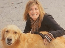 Cindy squats and poses with her arm around her golden retriever dog. She is smiling. She has shoulder length blonde hair and wears a brown jacket.