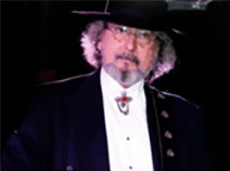 David plays bass guitar on stage while wearing a three piece suit. The suit jacket is black, but his vest and shirt are white. 