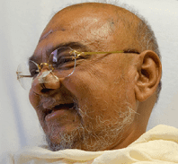 He smiles. He is bald. He wears glasses and has a gray wispy beard.