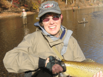 She wears a tan jacket, gray baseball cap, and sun glasses. She is holding a fish she caught.