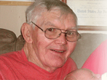He has gray hair, wears glasses, and a red t-shirt. He is holding an infant. He is smiling.