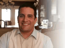 Eddie wears a striped white and tan shirt. He is tanned with short cropped brown hair.