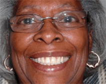 She has shoulder length gray hair. She is smiling brightly. She wears gold hoop earrings and silver glasses.