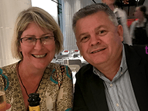 A couple sits at a restaraunt. She wears a gold shirt, has glasses, and has shoulder length blonde hair. He wears a gray striped shirt and black blazer jacket. He has gray hair.
