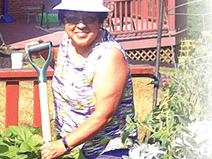 A woman stands in her garden holding a shovel. She wears a white bucket hat and a gray and white striped sleeveless blouse, with jeans.