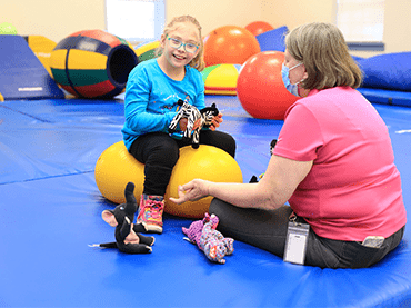Image of Tru on an exercise ball with her therapist holding a stuffed zebra. 