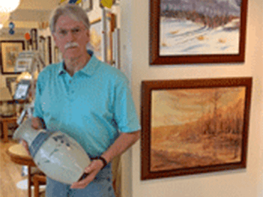 A man with gray hair and a gray moustache stands in a room next to a wall of artwork while holding a ceramic vase.