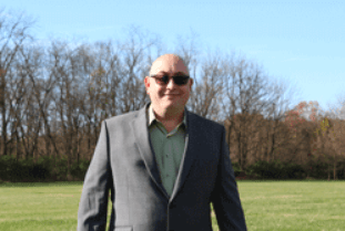 Dustin wears a charcoal gray suit and a white collared shirt. He has sunglasses. He is bald. He is standing outside in a field lined by trees.