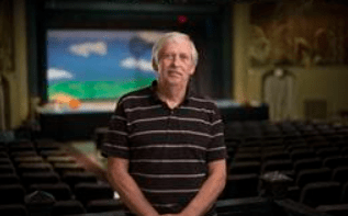 A man in a striped shirt is standing in a dimly lit room. He has gray hair.