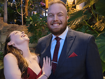 Dale is wearing a black tuxedo with a white collared shirt and red tie. He has a beard and red hair. Molly hugs him while laughing. She wears a red dress. She has long straight brown hair, half pulled back.